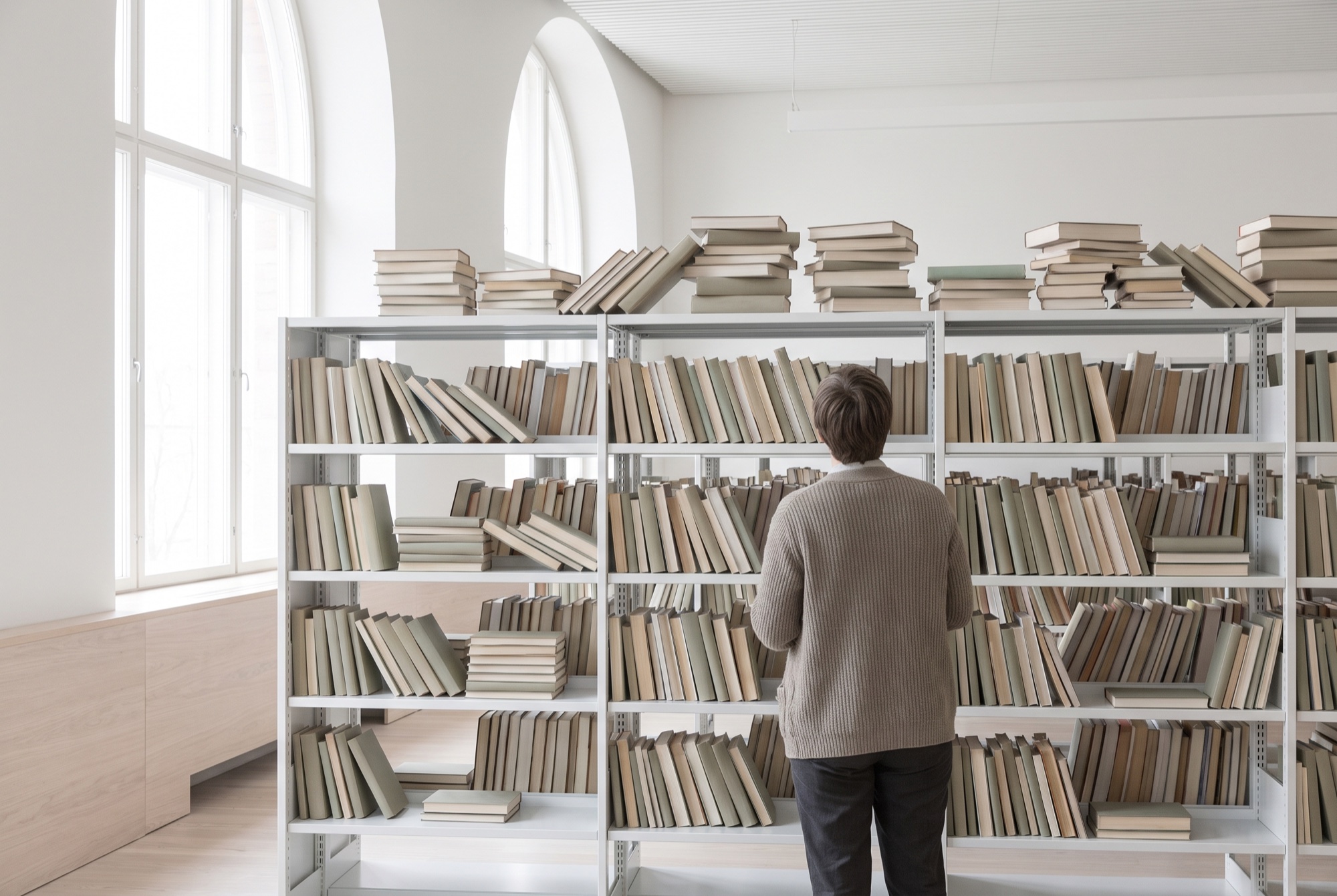 A librarian facing a run of overcrowded shelving, books stacked across the tops of the rows