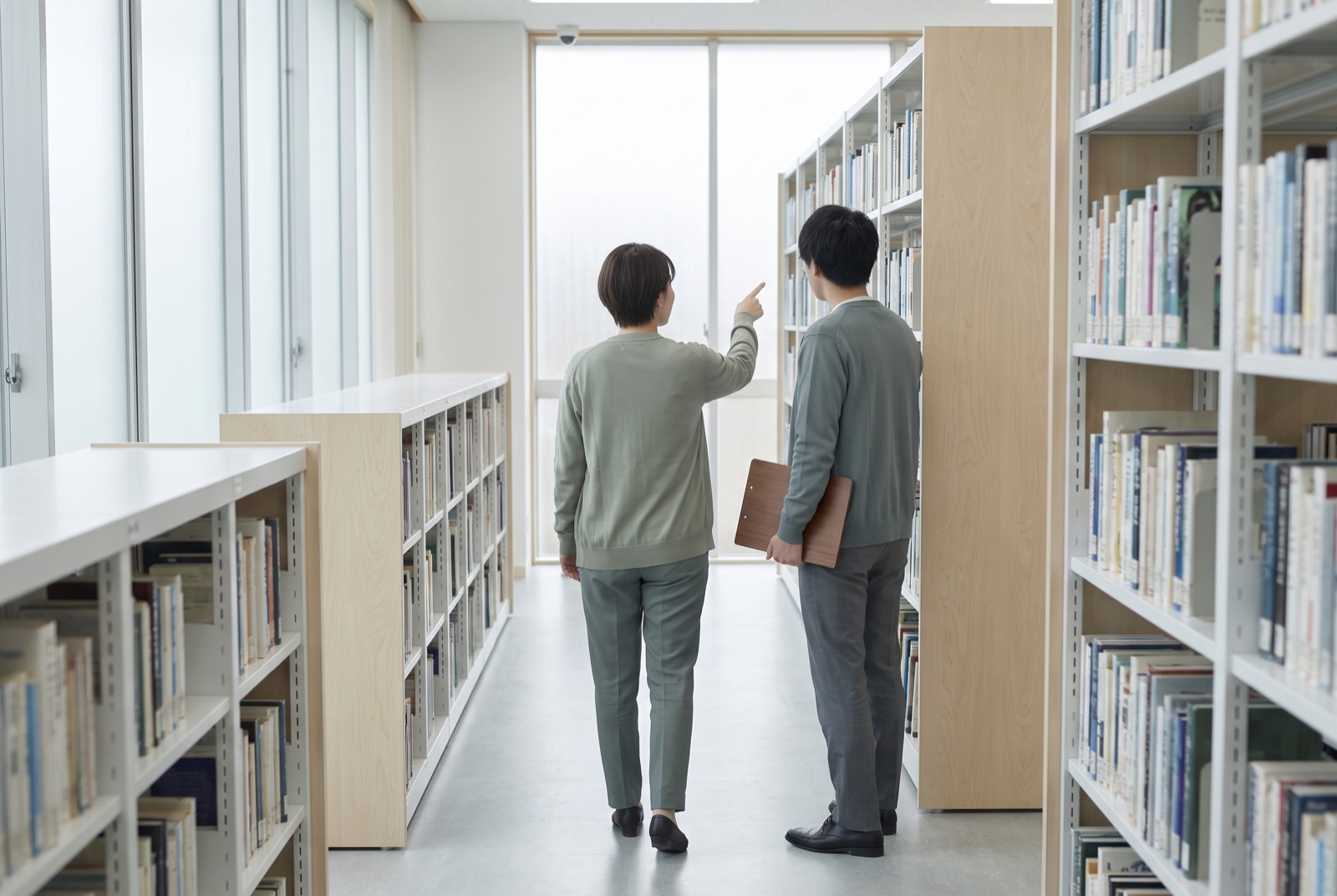 Two library colleagues reviewing a shelf section together along a bright aisle